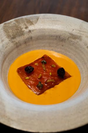 Elegant plating of orange carrot soup with dark garnishes and a rectangular protein floating in the center.