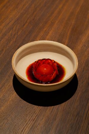 A bright red spherical dessert sits in a white bowl casting dramatic shadows on a wooden surface.