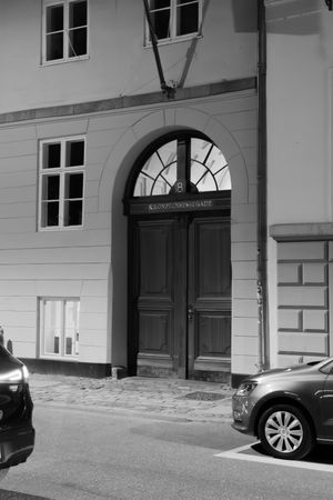 Black and white architectural view of an elegant building entrance with arched doorways and parked cars.