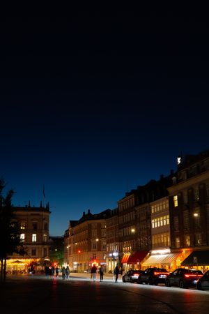 European city street at night with illuminated storefronts and car lights creating a warm glow against dark sky.