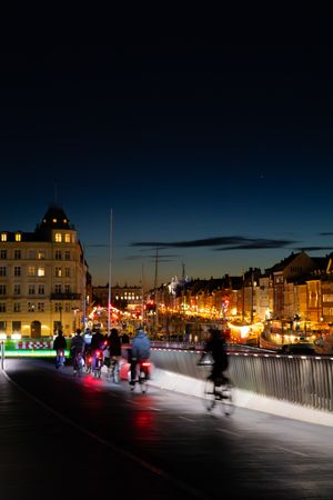 Waterfront cityscape at dusk with illuminated buildings and people walking along a lit promenade.
