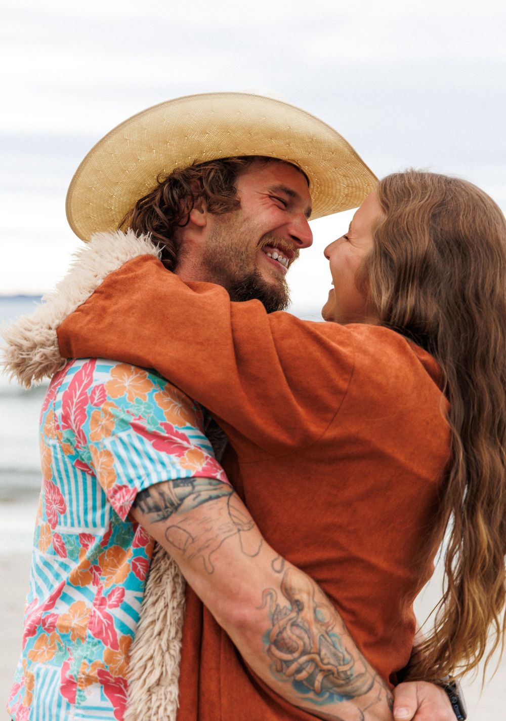 A couple embraces on the beach wearing a brown jacket and colorful floral print clothing at sunset.