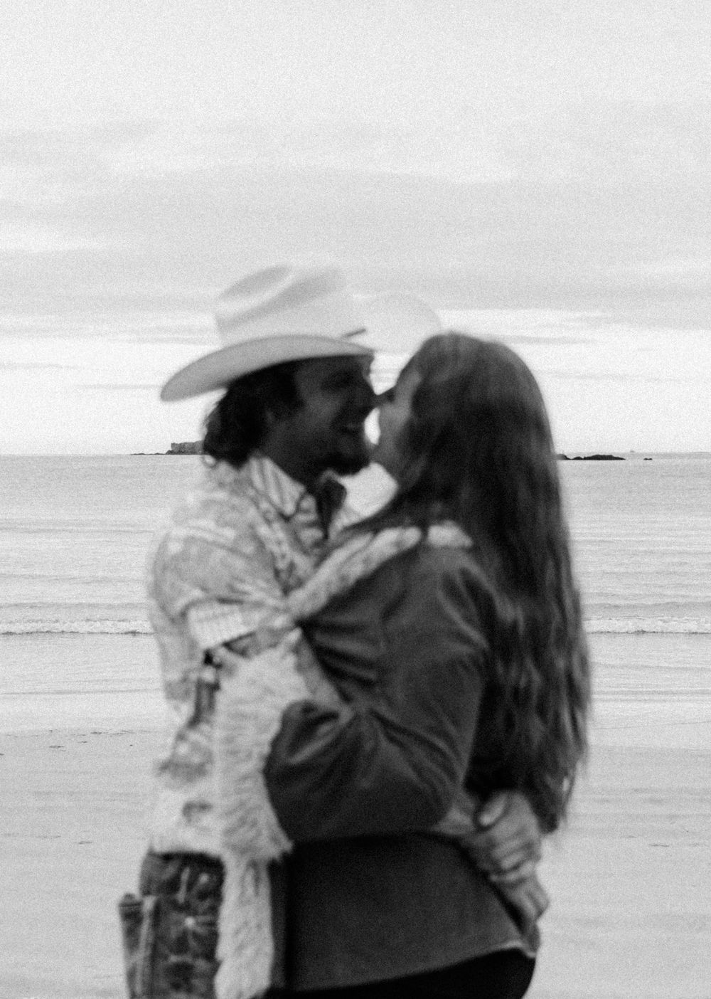 A romantic black and white photograph of a couple embracing on a beach with the ocean in the background.