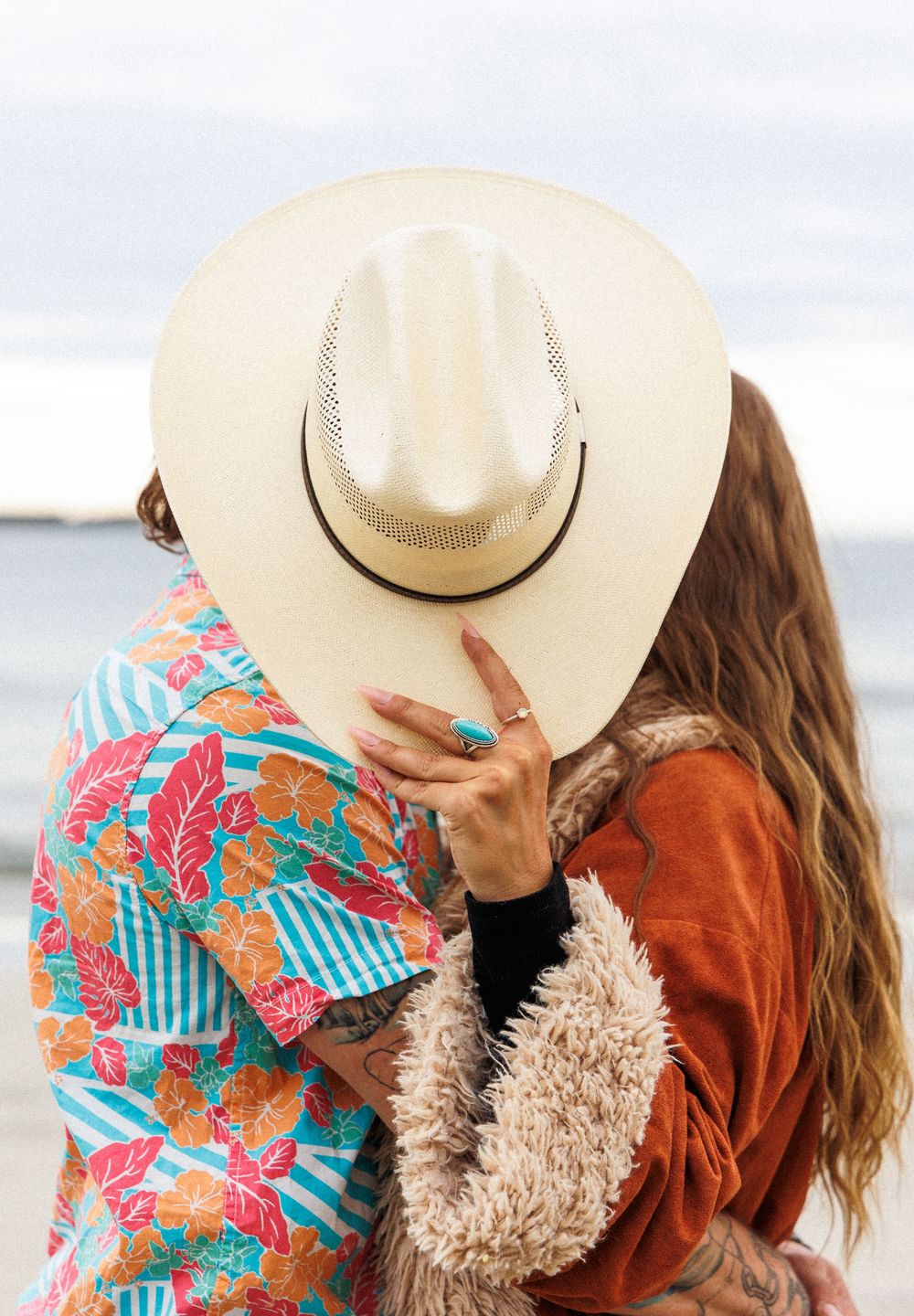 Two people in cozy sweaters and straw hats share tender moments together at the beach.