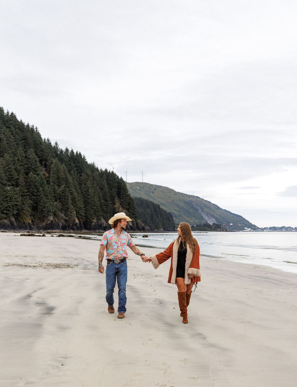 A couple in casual clothing holds hands while taking a romantic walk along a misty beach with evergreen trees visible.