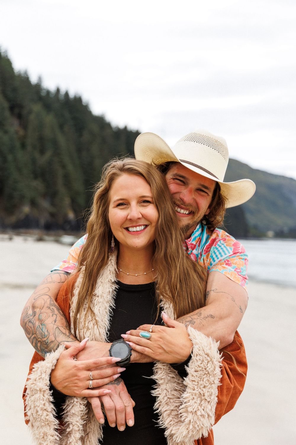 A romantic couple in western attire and a fuzzy jacket embrace and laugh together on a scenic beach.
