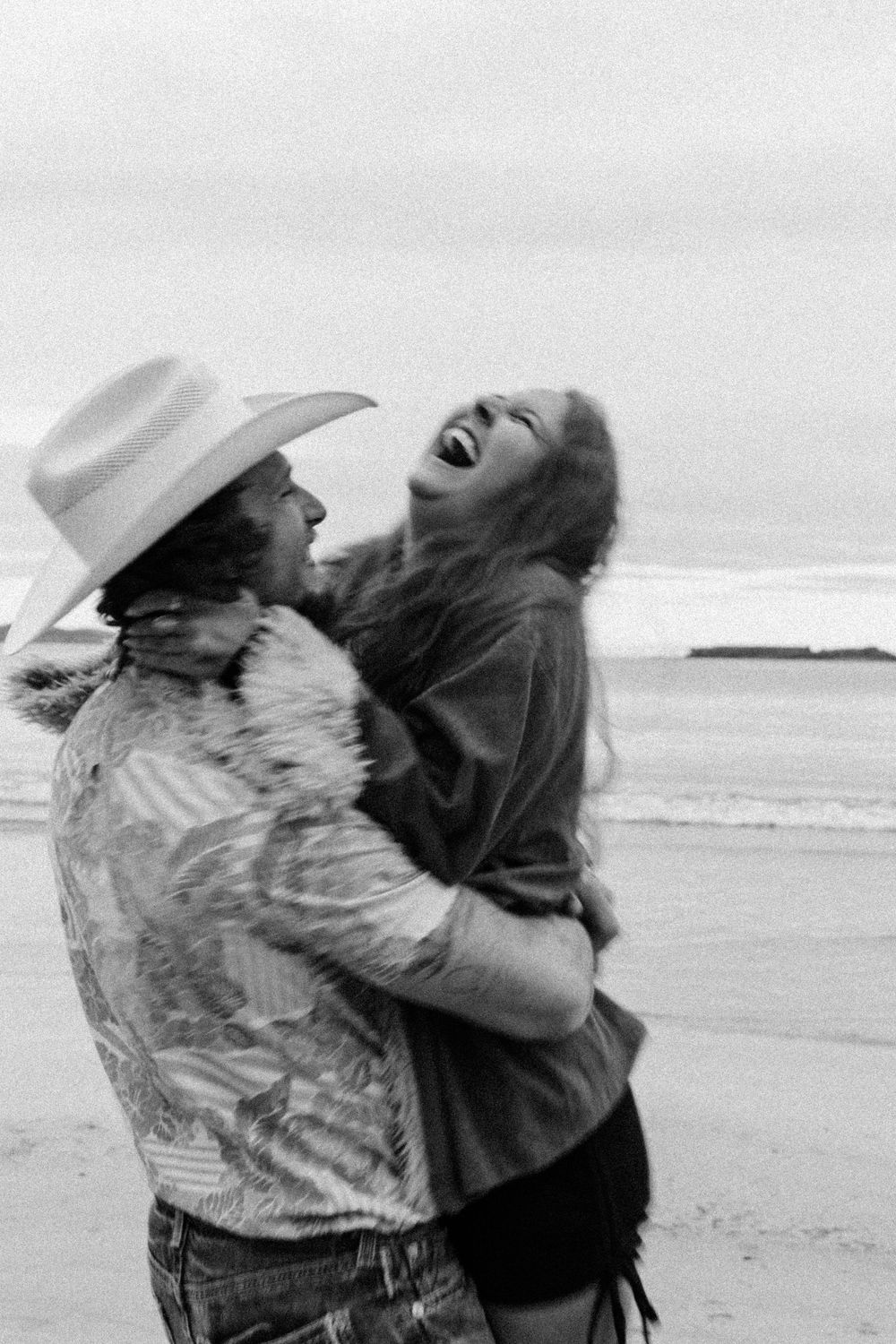 Two people share a joyous embrace and laugh together on a breezy beach in black and white photography.