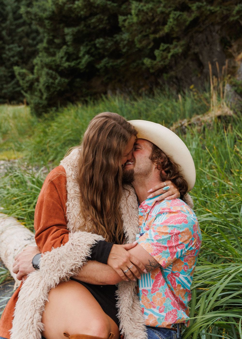 A romantic moment between a couple sitting in tall grass with a person wearing a cowboy hat and colorful tropical shirt.