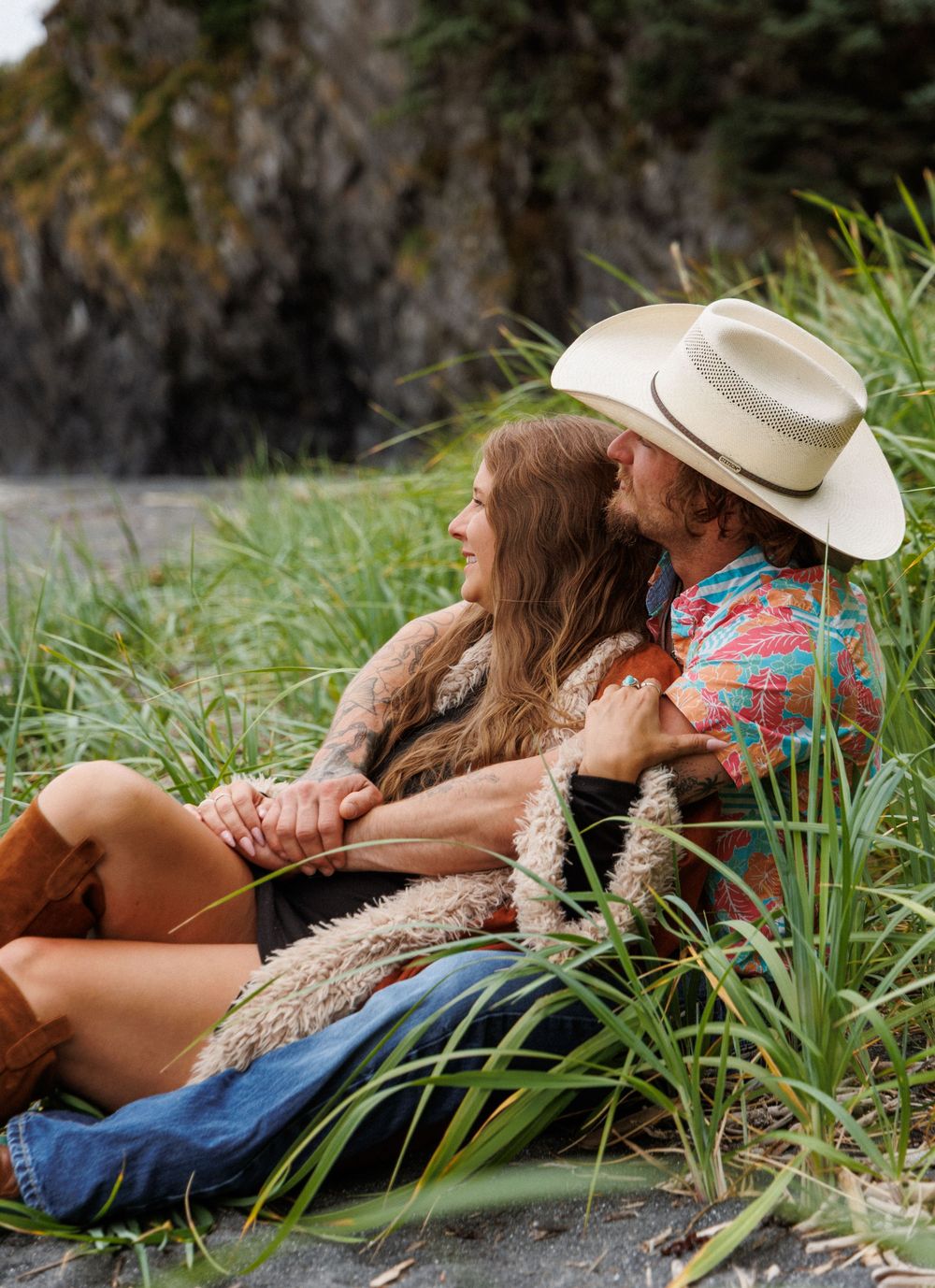 A couple in cowboy boots and western attire relax together on grassy coastal terrain.