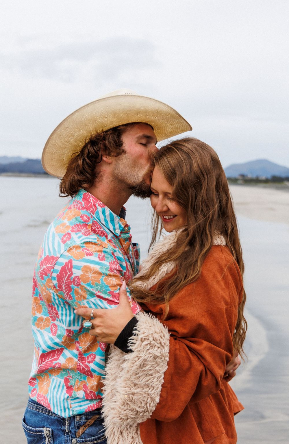 A couple in western attire shares a romantic moment on a beach with mountains visible in the background.