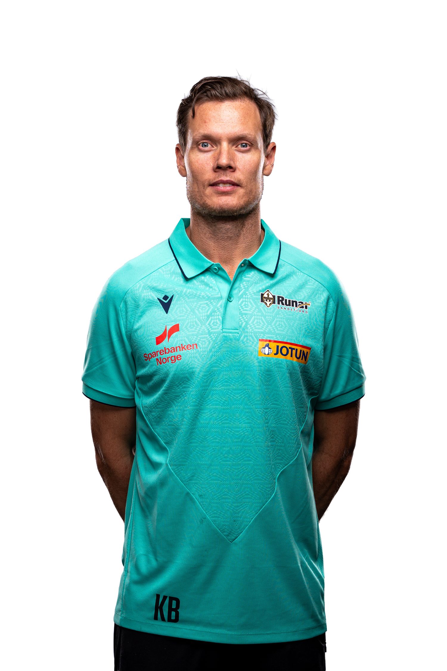 Soccer referee wearing a turquoise polo shirt with sponsorship logos and official patches stands against white background.