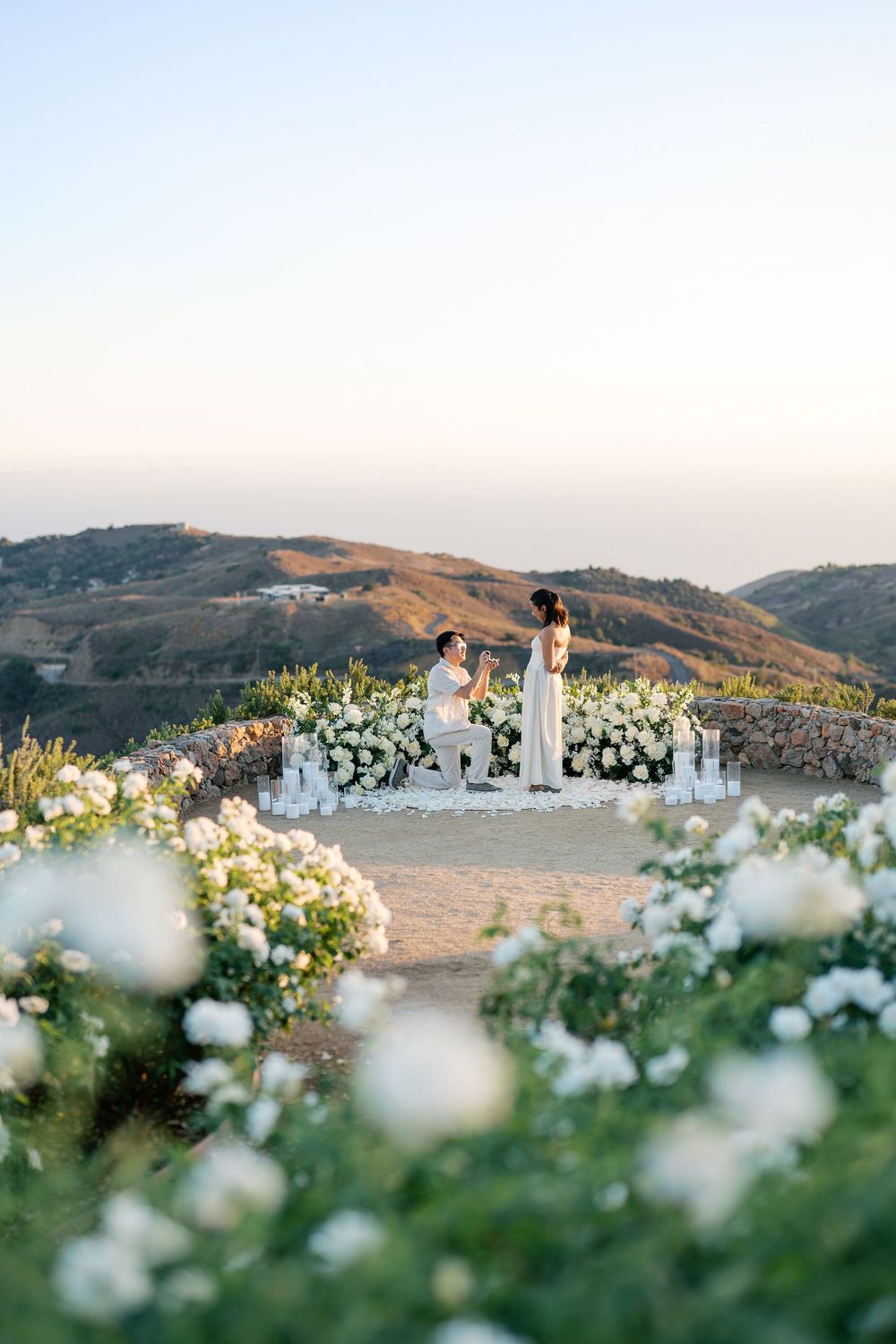 Scenic outdoor wedding ceremony setup with white flowers and mountain landscape views at sunset.