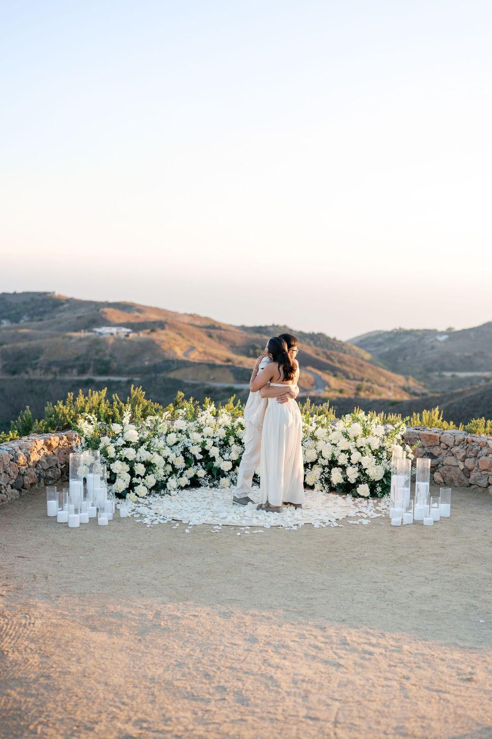 A romantic mountain-top proposal scene with white floral arrangements and candles at golden hour against rolling hills.