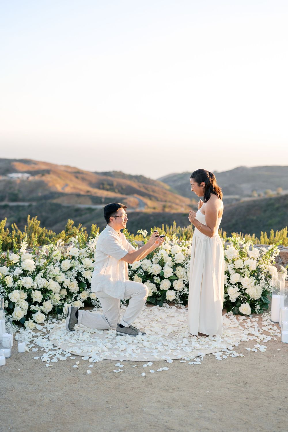 A romantic beach proposal with white flowers scattered on the sand at sunset with beautiful mountains in the background.