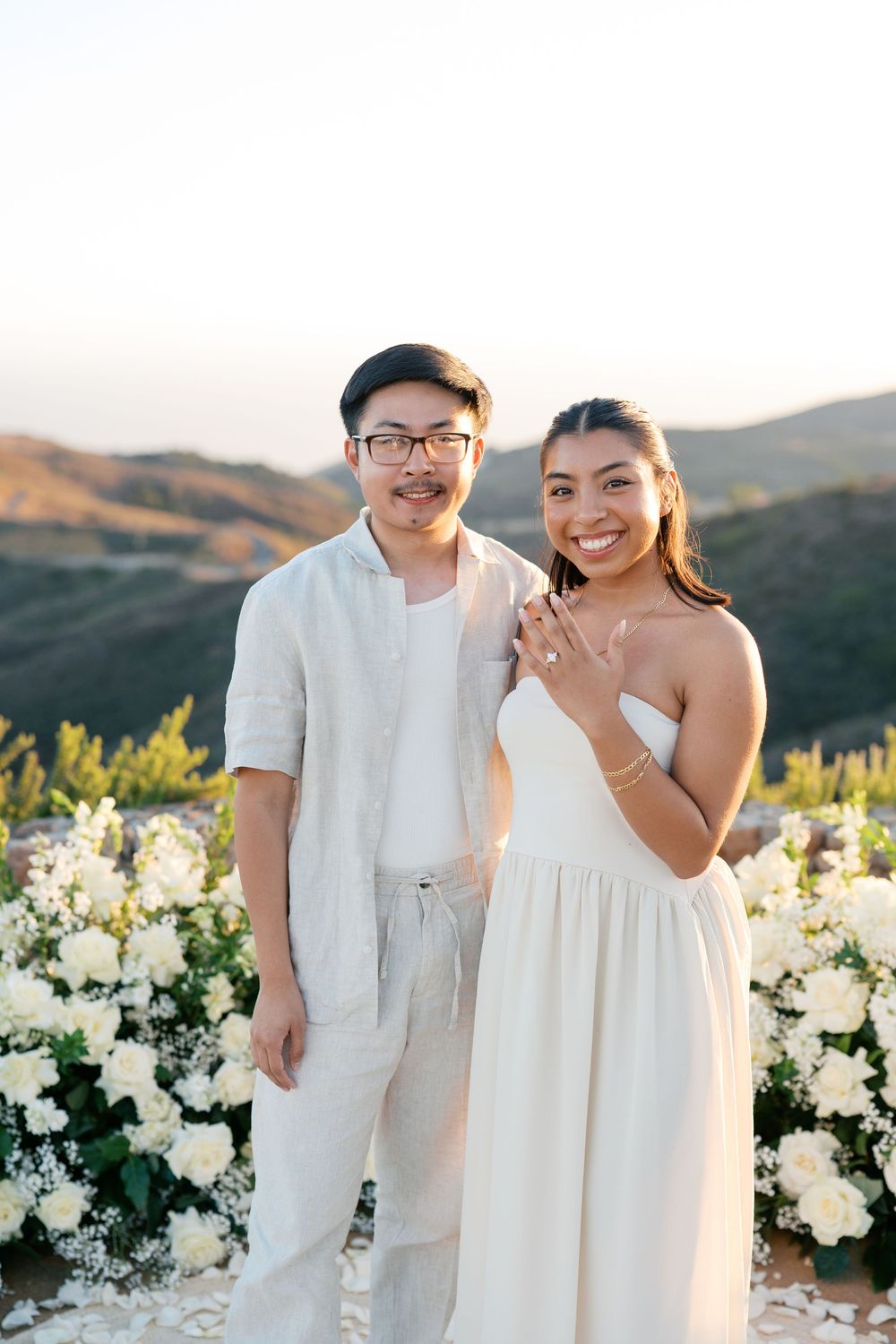 A couple in white outfits poses together against a scenic mountain backdrop with white floral decorations at sunset.