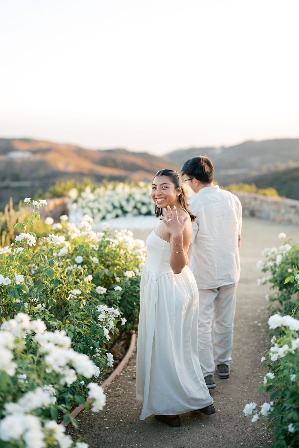 Romantic moment captured in a sunlit garden path lined with white roses and scenic mountain views.