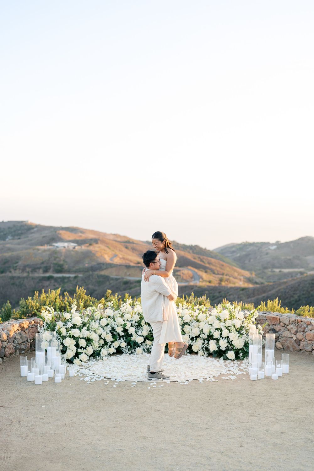 A romantic outdoor wedding scene at sunset with white floral arrangements and glowing candles on a stone patio.