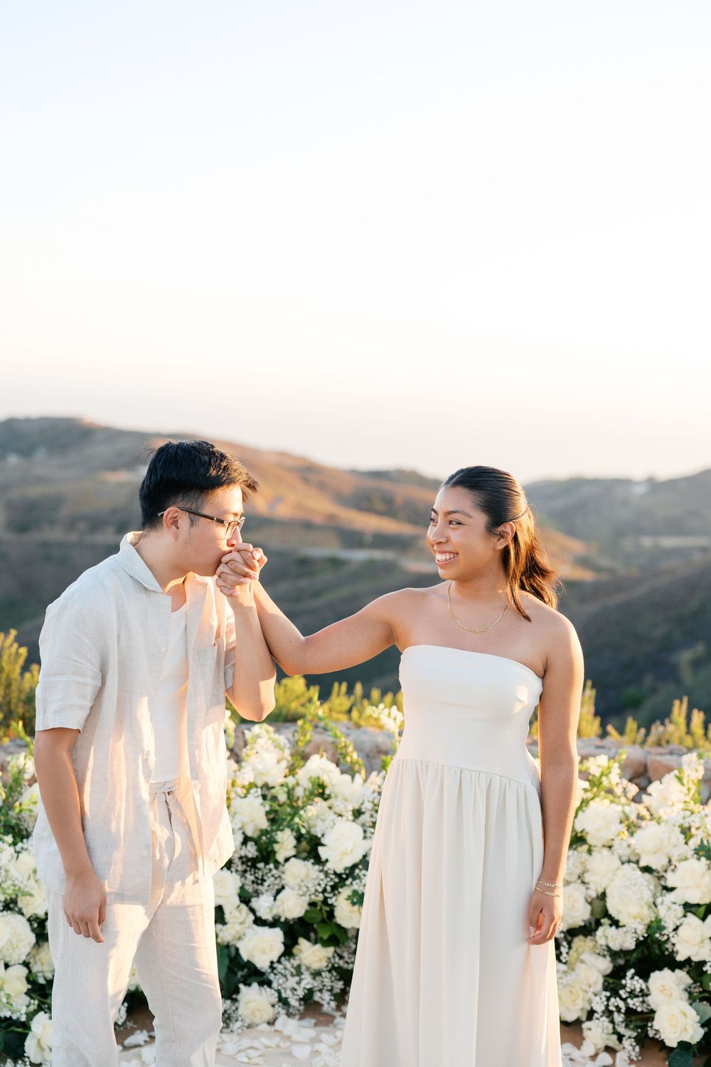 A romantic couple stands together in an outdoor garden with white flowers and mountains in the background at sunset.