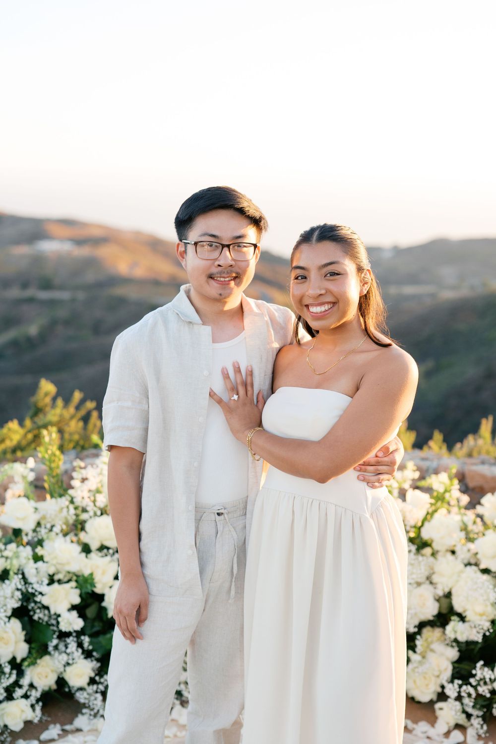 A couple in white outfits embraces during an outdoor sunset engagement photo session surrounded by white flowers.