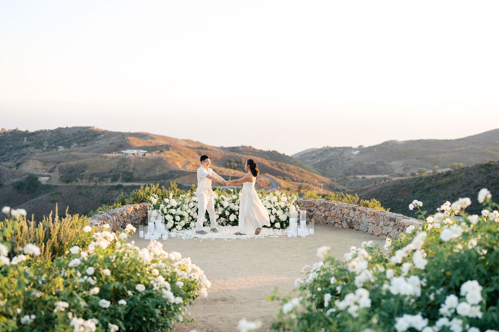 A scenic outdoor wedding ceremony setup on a mountaintop terrace surrounded by blooming white roses and wildflowers.