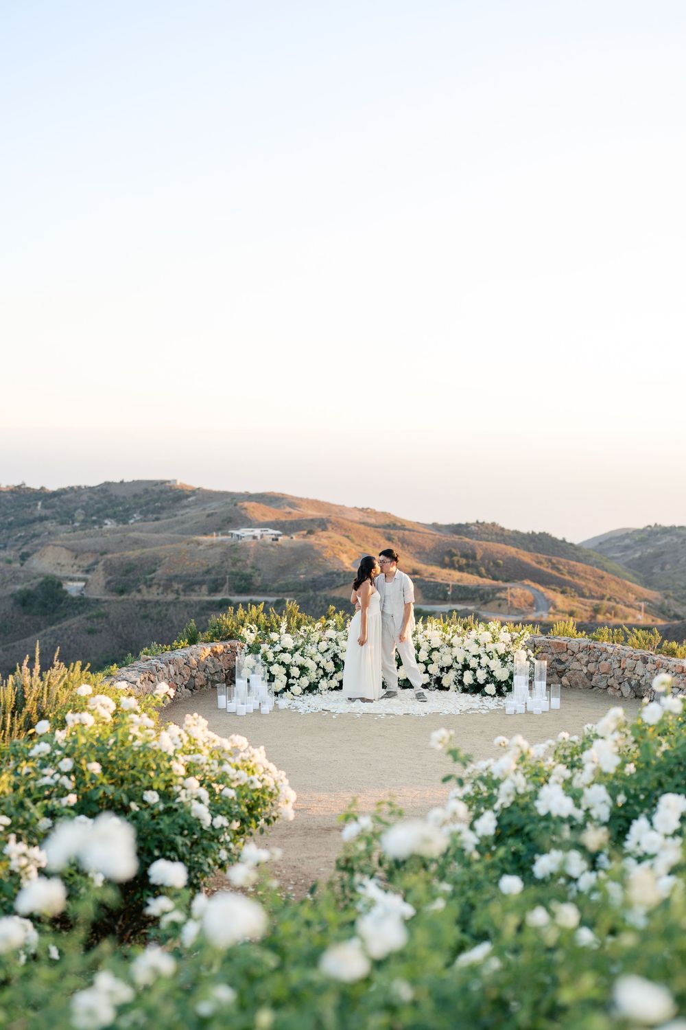 Sunset outdoor wedding ceremony setup overlooking rolling hills with white floral arrangements and standing guests.