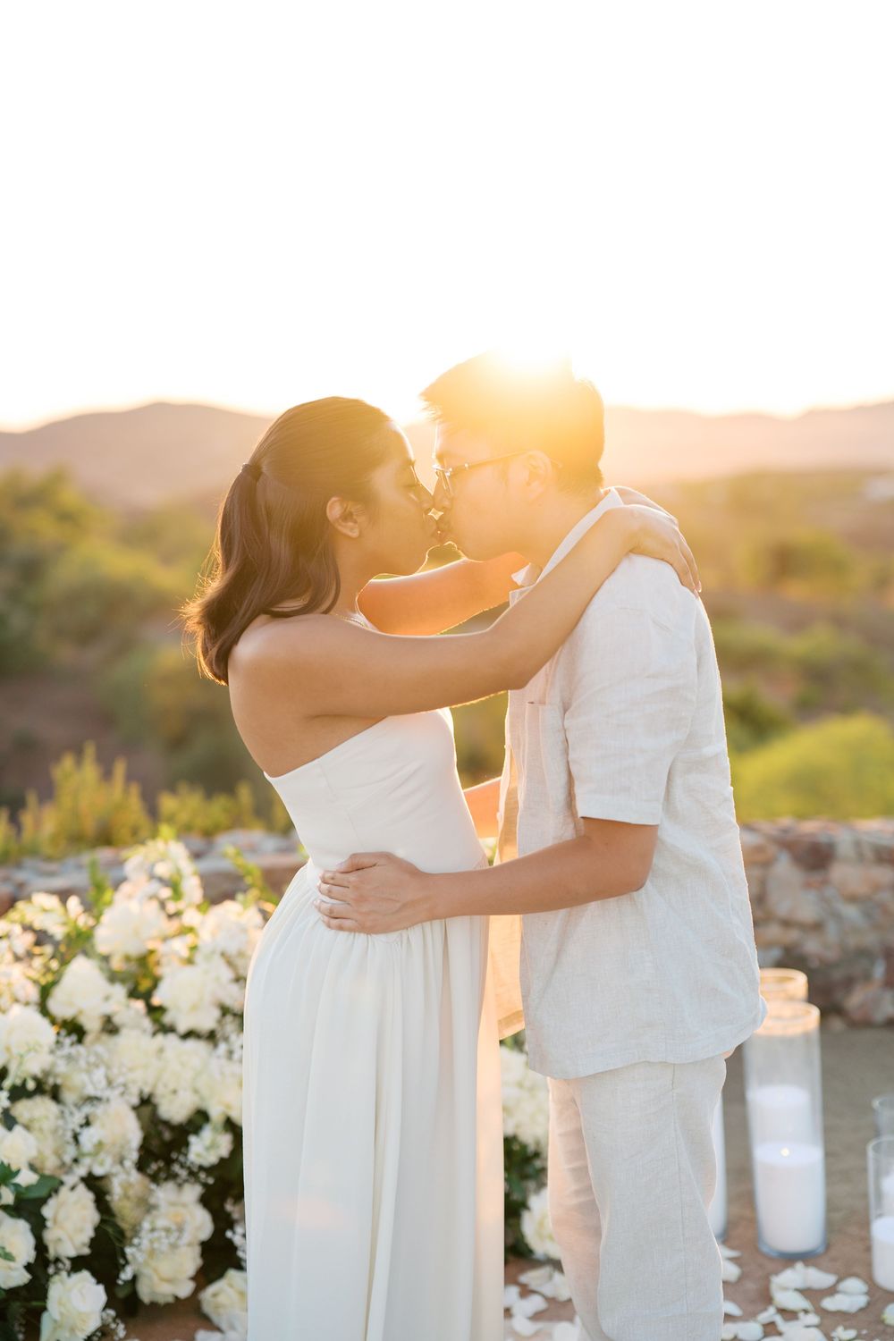 Couple in white embracing during golden hour at sundown with white floral decorations in background.