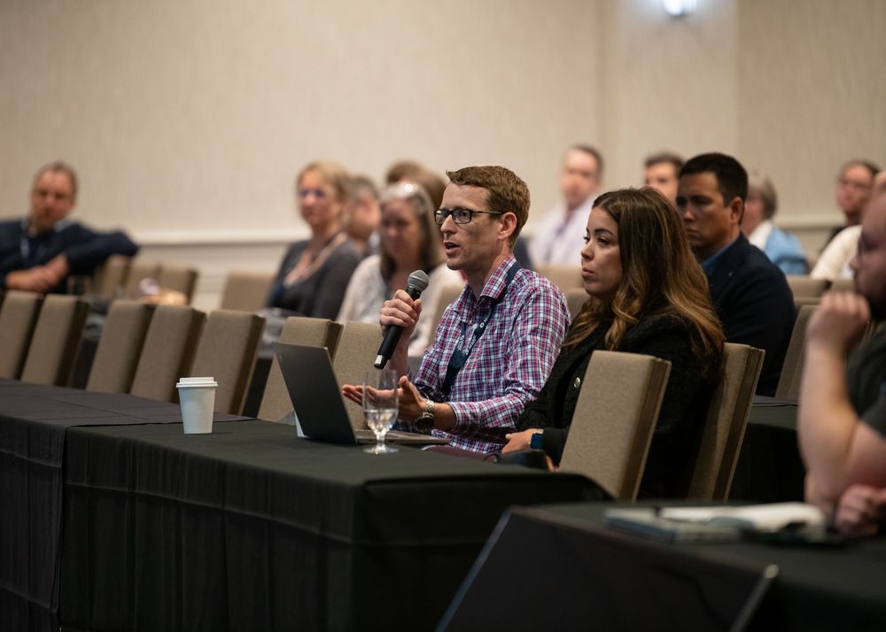Professionals at a conference panel discussion, man speaking into microphone with laptop open at table.