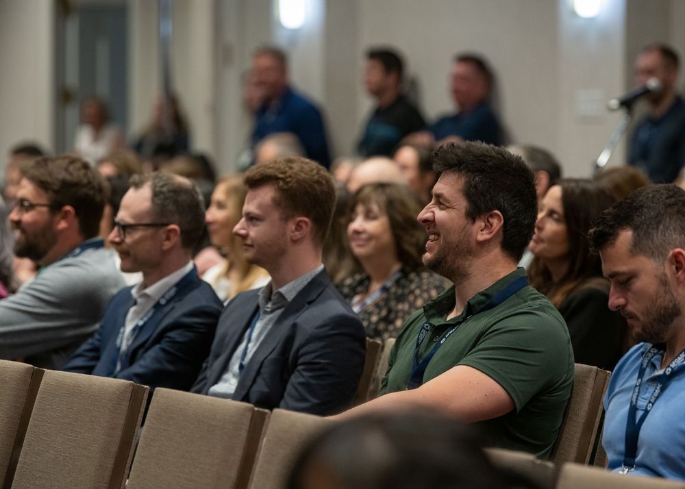 Audience members smile and laugh while seated in rows at an indoor conference or corporate event.