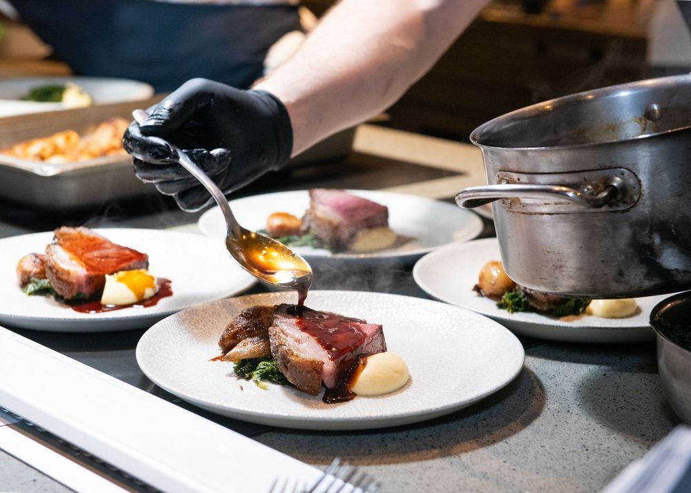 Chef plating a seared meat dish and spooning sauce over it in a restaurant kitchen.