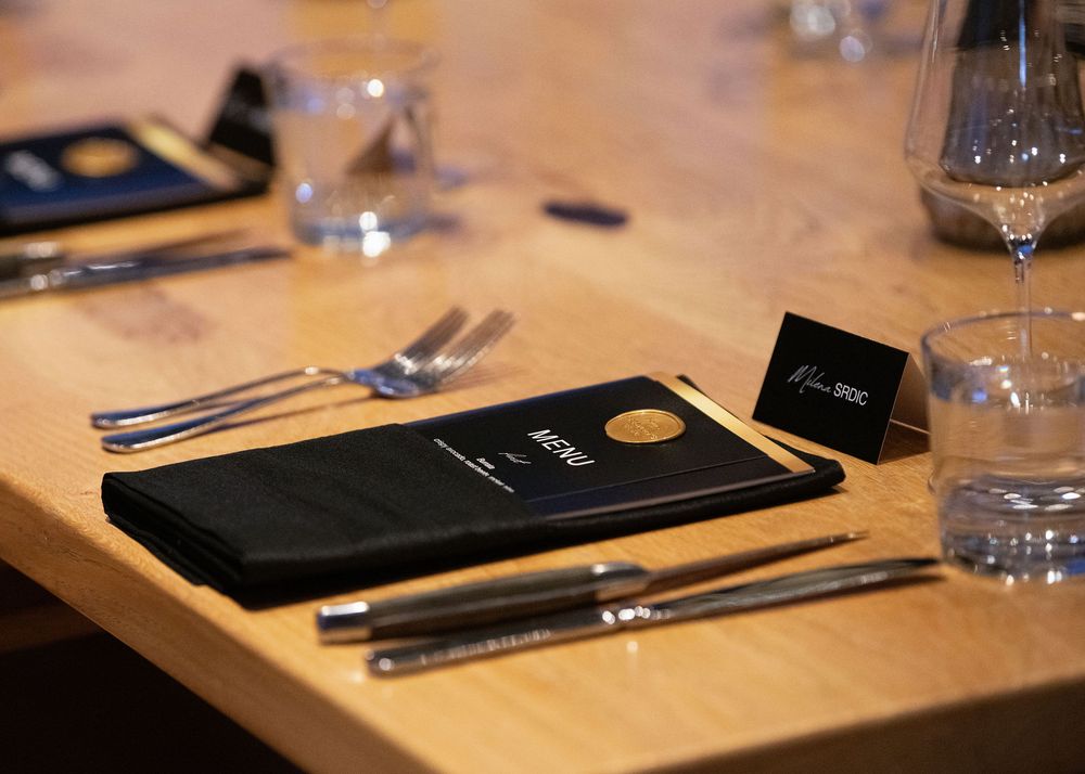 Formal place setting with black menu card, cutlery, and water glass on a wooden banquet table.