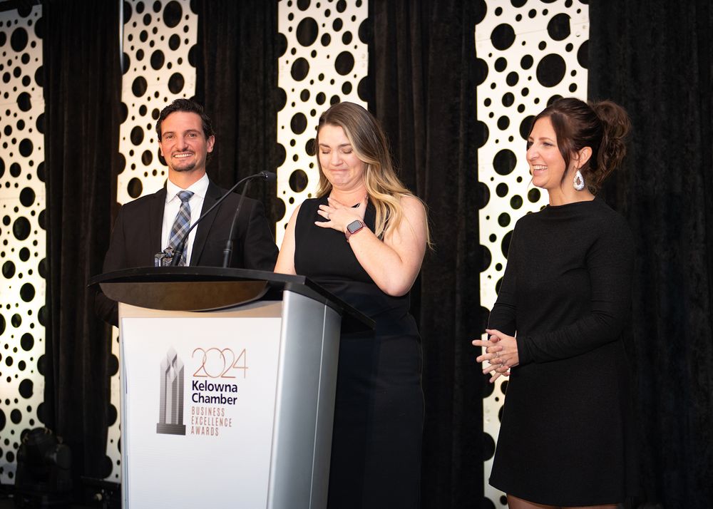 Woman in black dress smiling with hand on heart while receiving award on stage at Kelowna Chamber Business Excellence Awards.