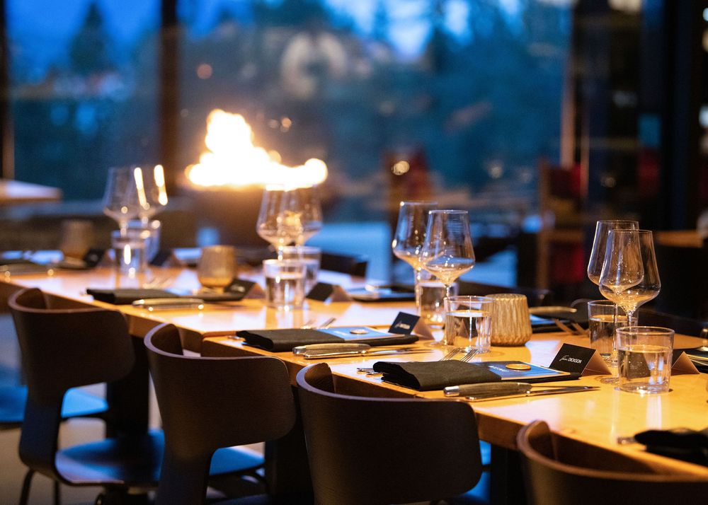Elegantly set dining table with wine glasses and candles beside a glowing outdoor fire feature at dusk.