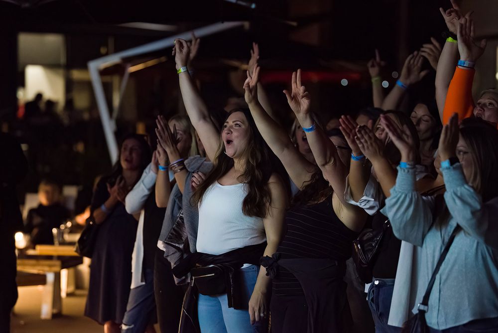 Excited crowd of women raising hands at an indoor concert or event in a dimly lit venue.