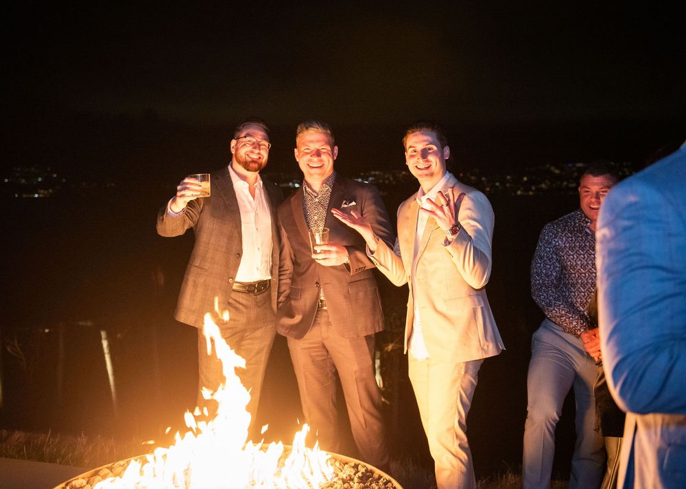 Three men in suits standing around an outdoor fire pit at night, holding drinks and smiling at the camera.