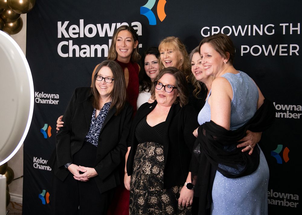 Seven women smiling in front of Kelowna Chamber backdrop at the Business Excellence Awards - event photo by Tania Veitch.