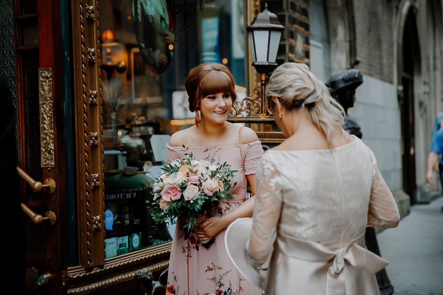 Bride and her wedding party walking down a narrow European street in formal attire with pink floral bouquets.