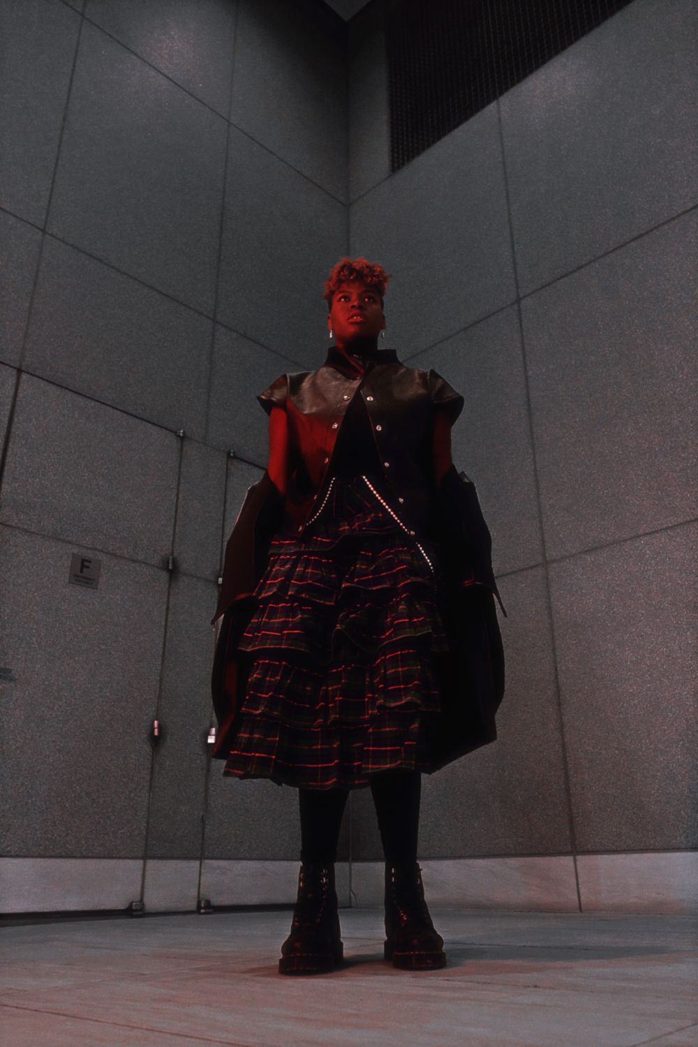 Two figures in black and red outfits stand in a dimly lit concrete room in a moody cinematic scene.