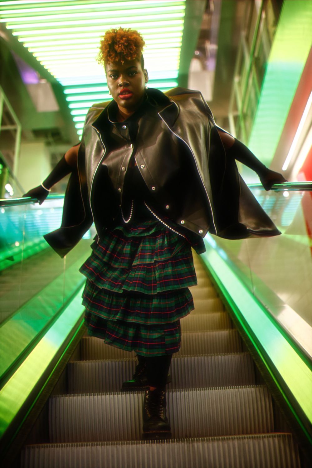 A figure in a plaid ruffled skirt and black leather jacket poses confidently on a green-lit escalator.
