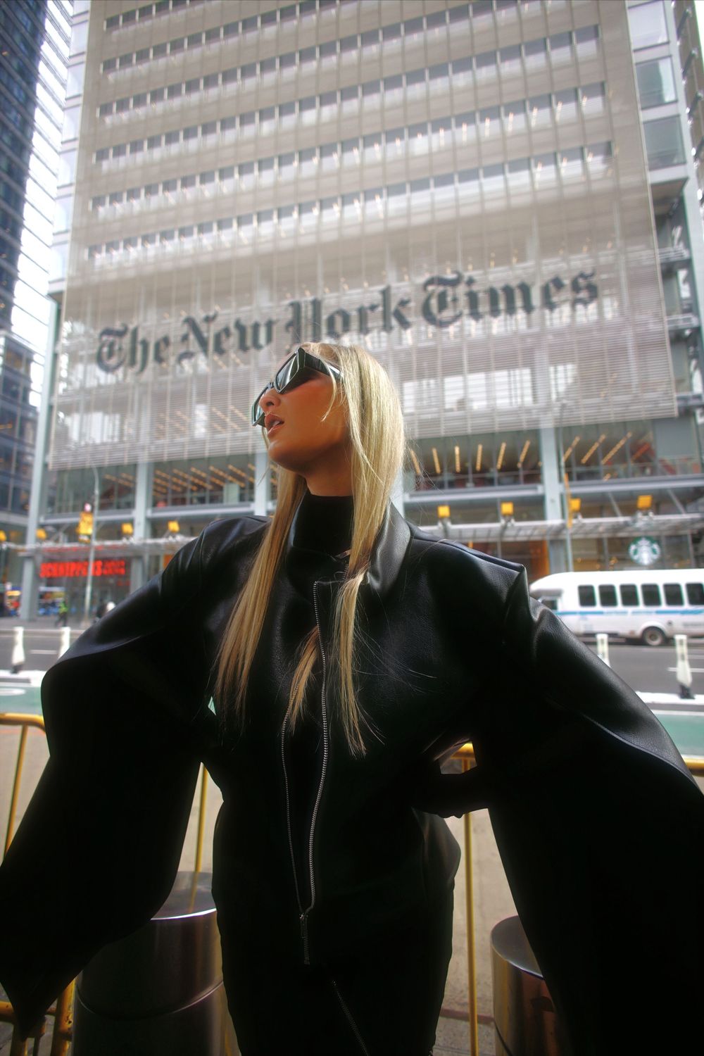 A person in a black top stands with arms outstretched in front of the New York Times building.