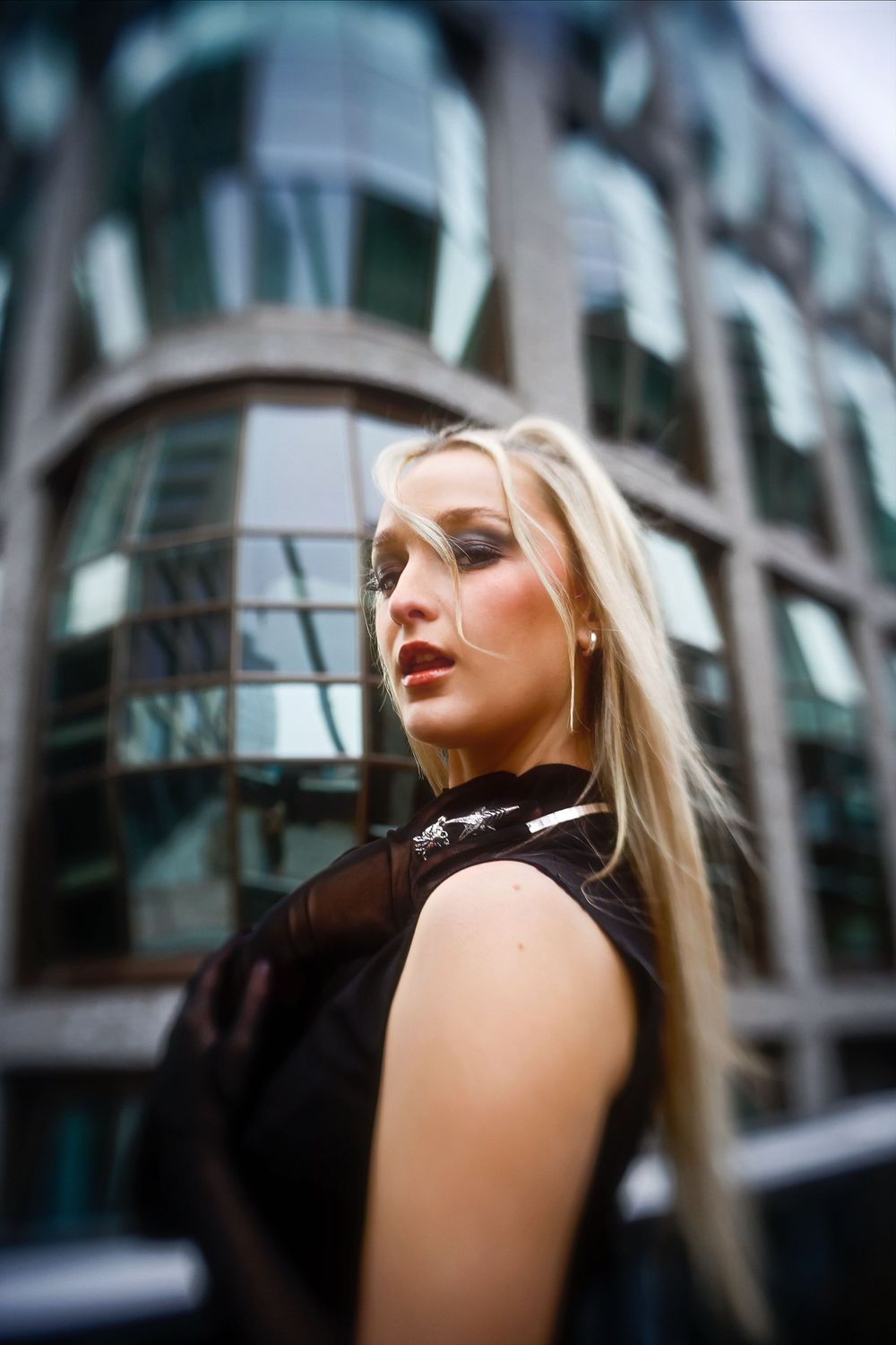 A fashion portrait against a modern glass building features sleek black attire and dramatic lighting.