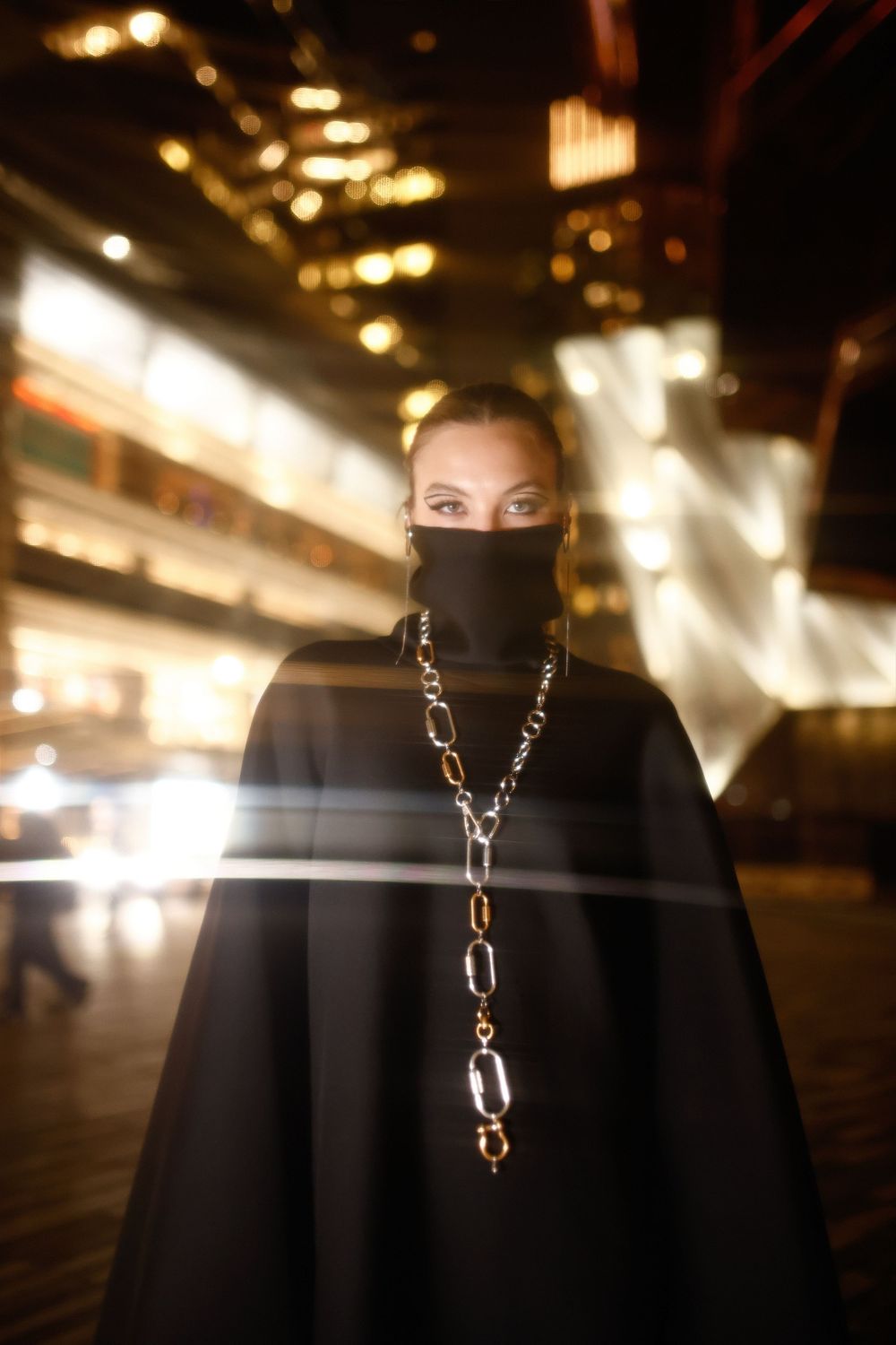 Dramatic nighttime portrait with chain necklace and dark clothing against blurred city lights in background.