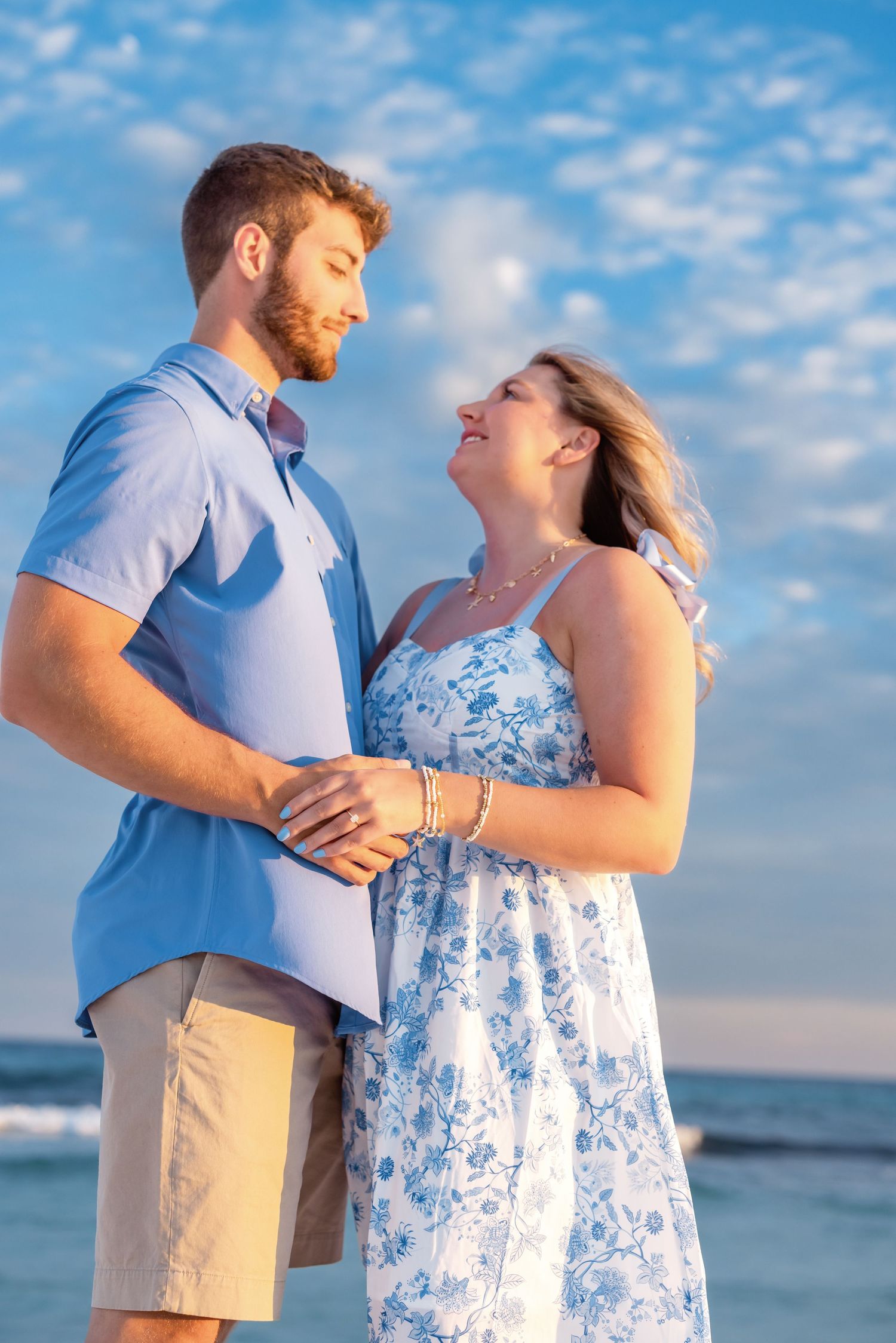 A couple in beach attire sharing a tender moment by the ocean as the sun sets behind fluffy clouds.