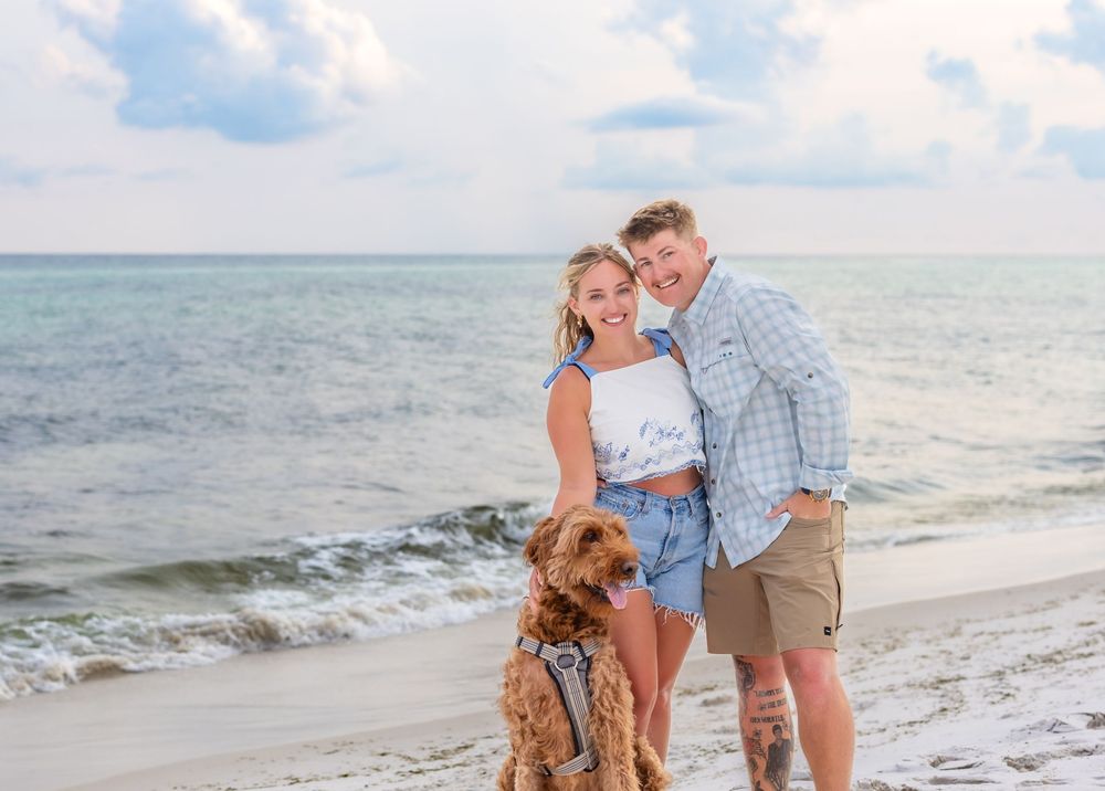 A couple stands on the beach with their golden doodle dog while waves crash on the shore under a cloudy sky.