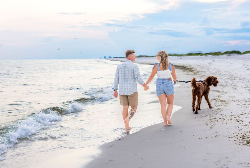 A couple walks their brown dog along a serene beach shoreline at sunset.