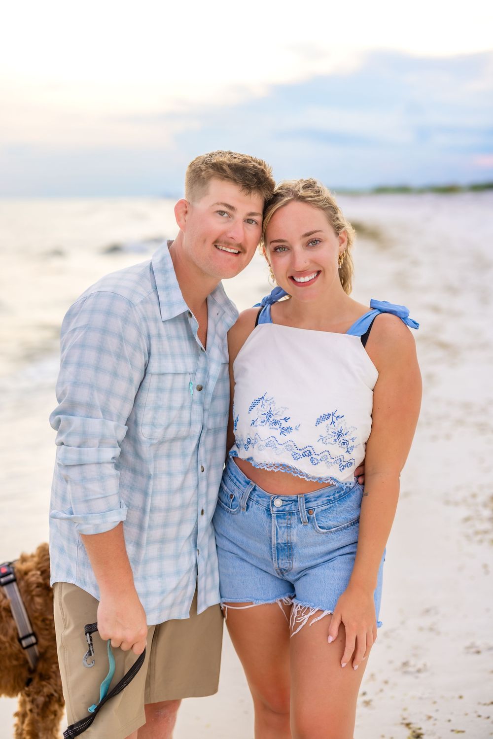 A young couple smiles while enjoying a romantic sunset walk together on a white sandy beach.