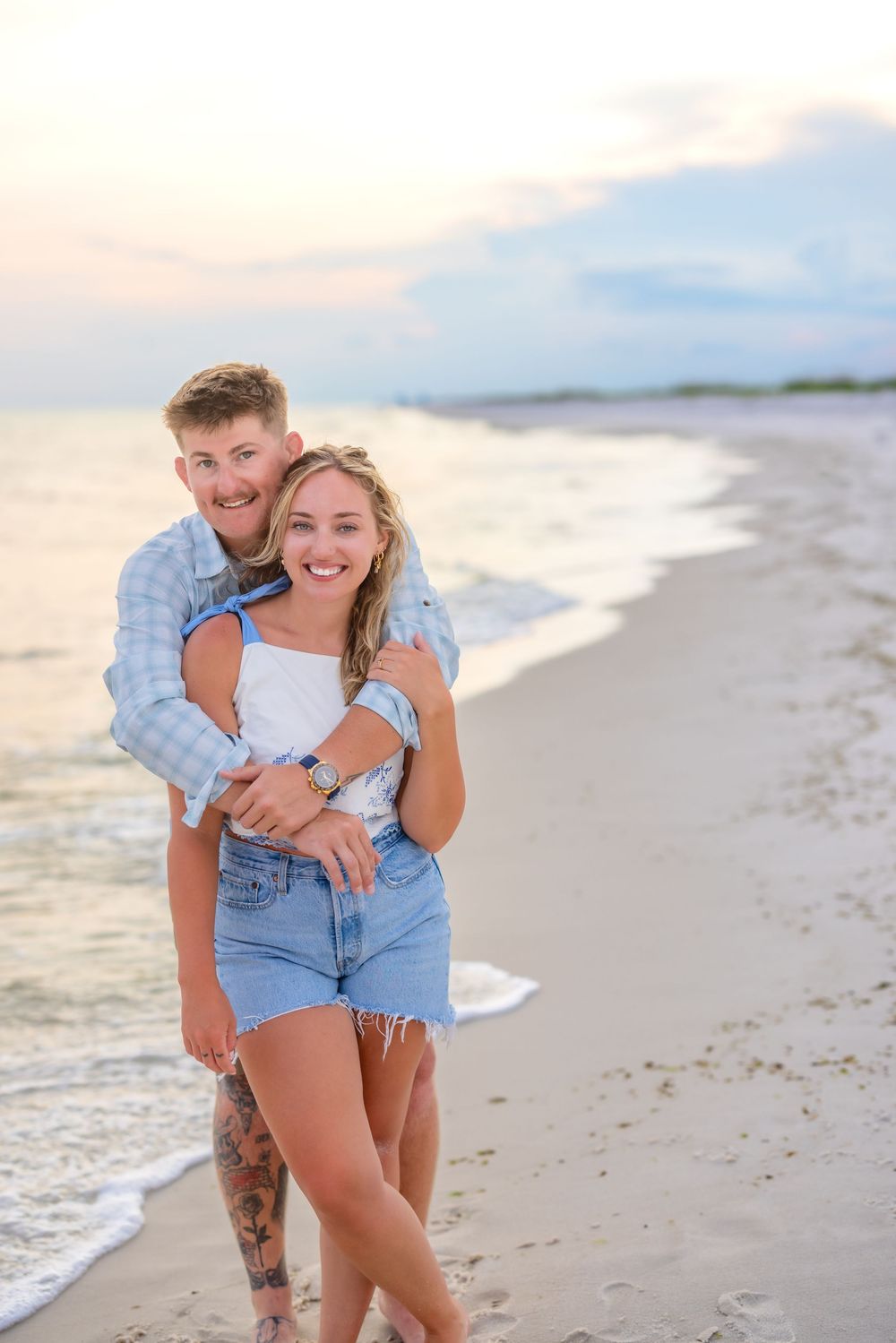 A couple embraces playfully on a scenic beach at sunset with waves crashing behind them.