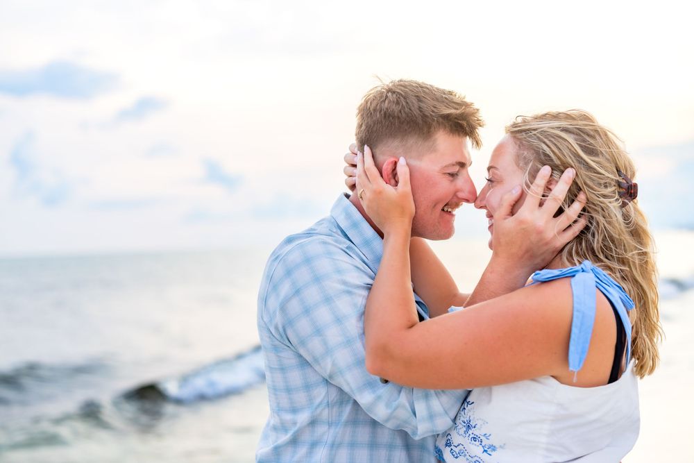 A couple in blue and white outfits shares romantic kisses along a scenic beach during golden hour.