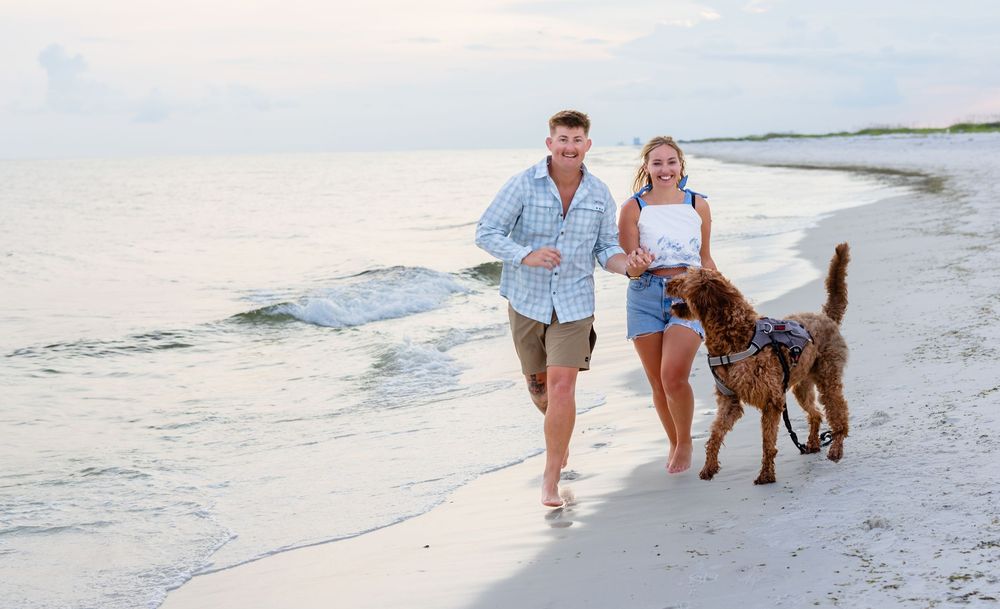 A couple and their dog enjoy a playful walk along a sandy beach at sunset with waves rolling in.