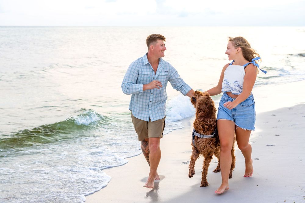 Couple walking their golden brown dog along a sunny beach shoreline while waves crash behind them.