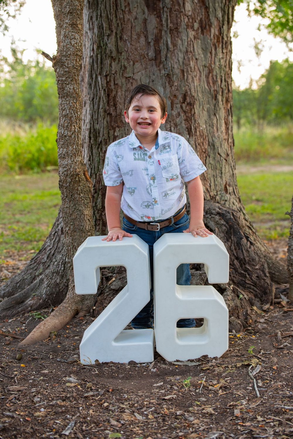 Child sitting playfully with decorative white number 26 letters against tree trunk in outdoor setting.
